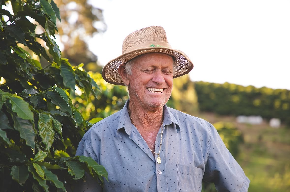Luiz Carlos Zanette wearing a straw hat and a light blue button-up shirt stands outdoors in a sunny farm setting with green plants in the foreground and a blurred landscape behind him.