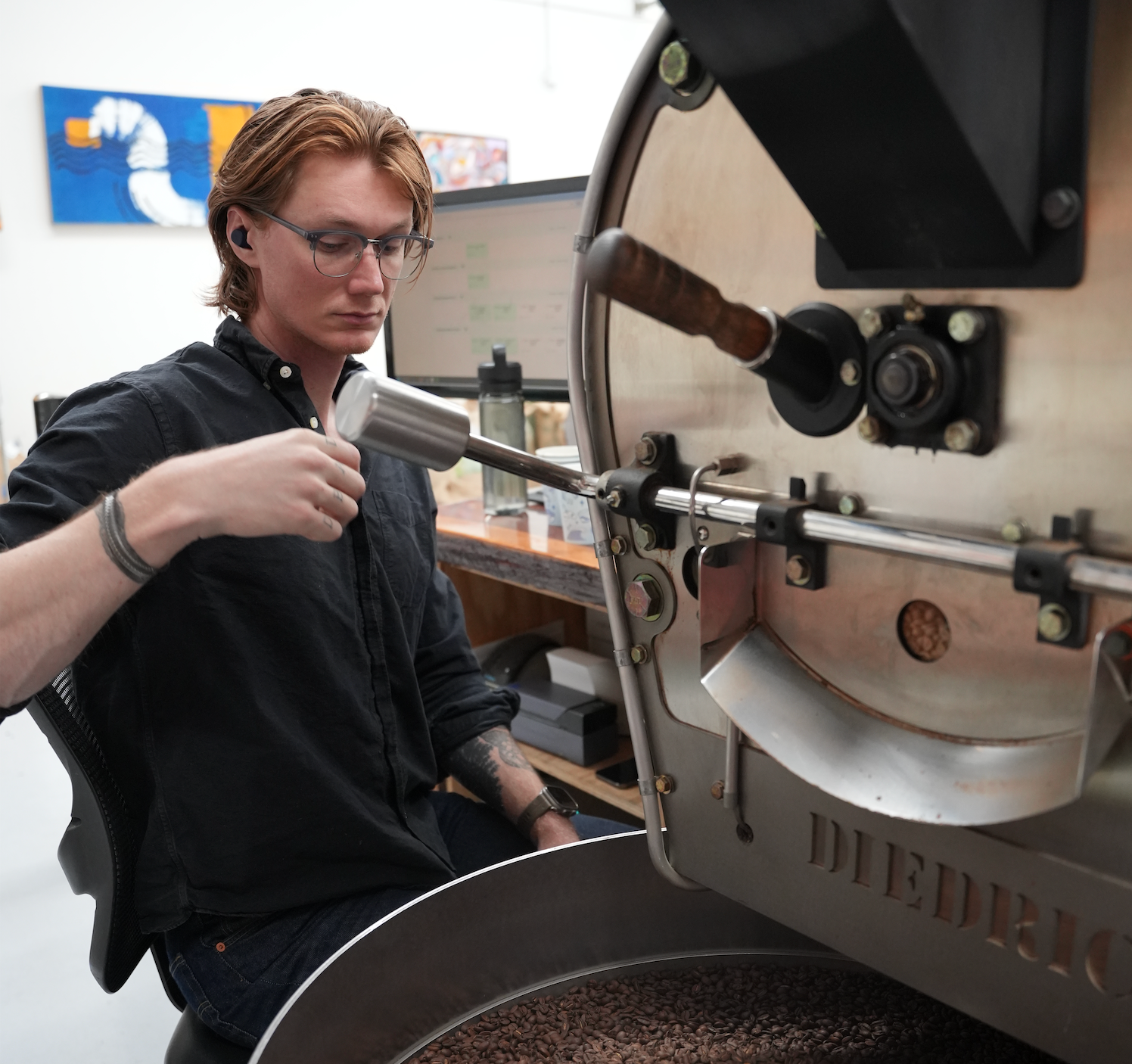 A man in a black shirt operates a large stainless steel coffee roaster, using a metal tool inside the chamber. The word 'DIEDRICH' is visible on the front of the machine, and roasted coffee beans are visible in the hopper below.